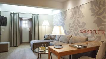 Quarto de hotel com sofá, mesa com abajur, mesinha lateral, televisão e janela com cortinas.