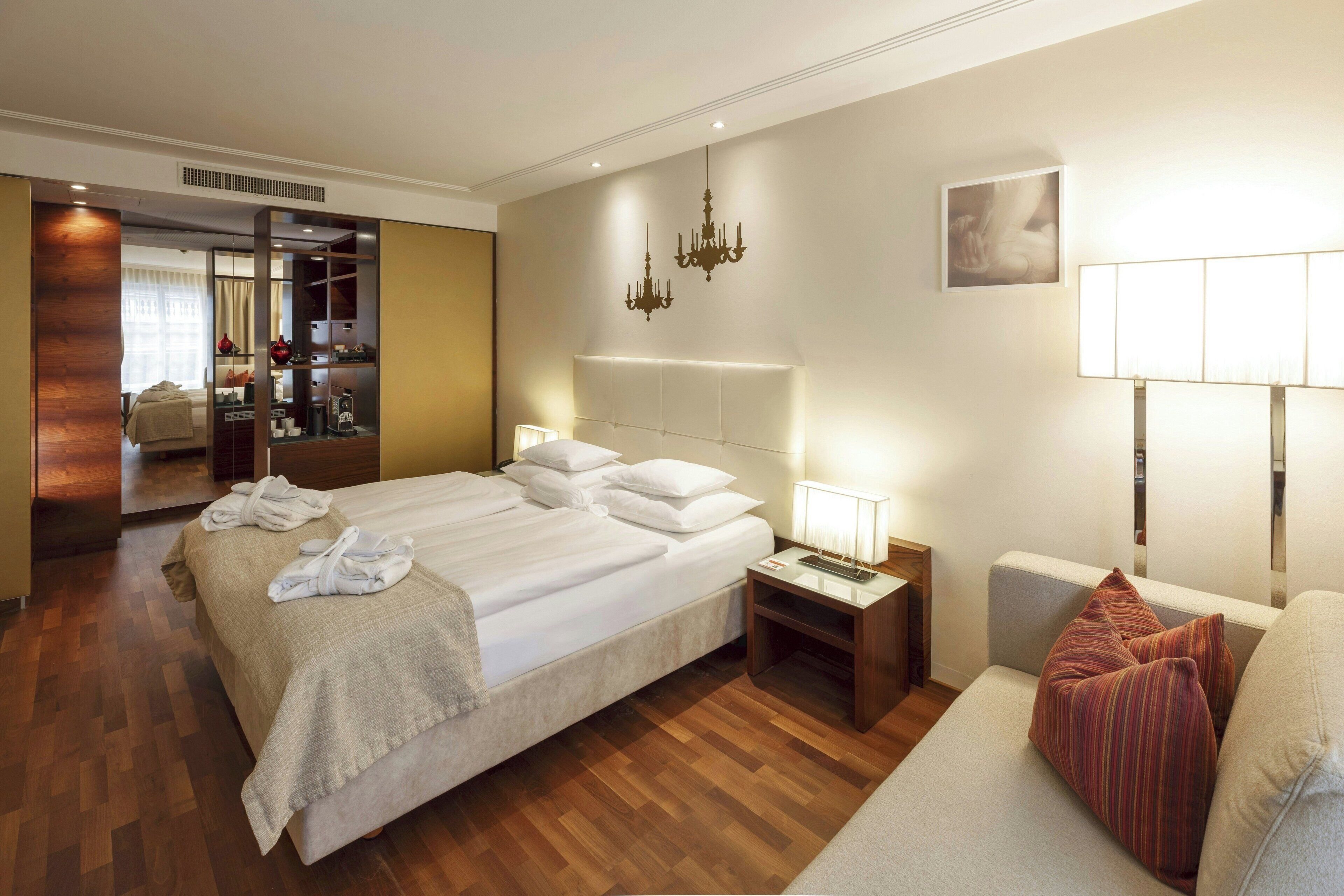 A hotel room with a large bed, bedside tables, a sofa, a mirror, and a chandelier.