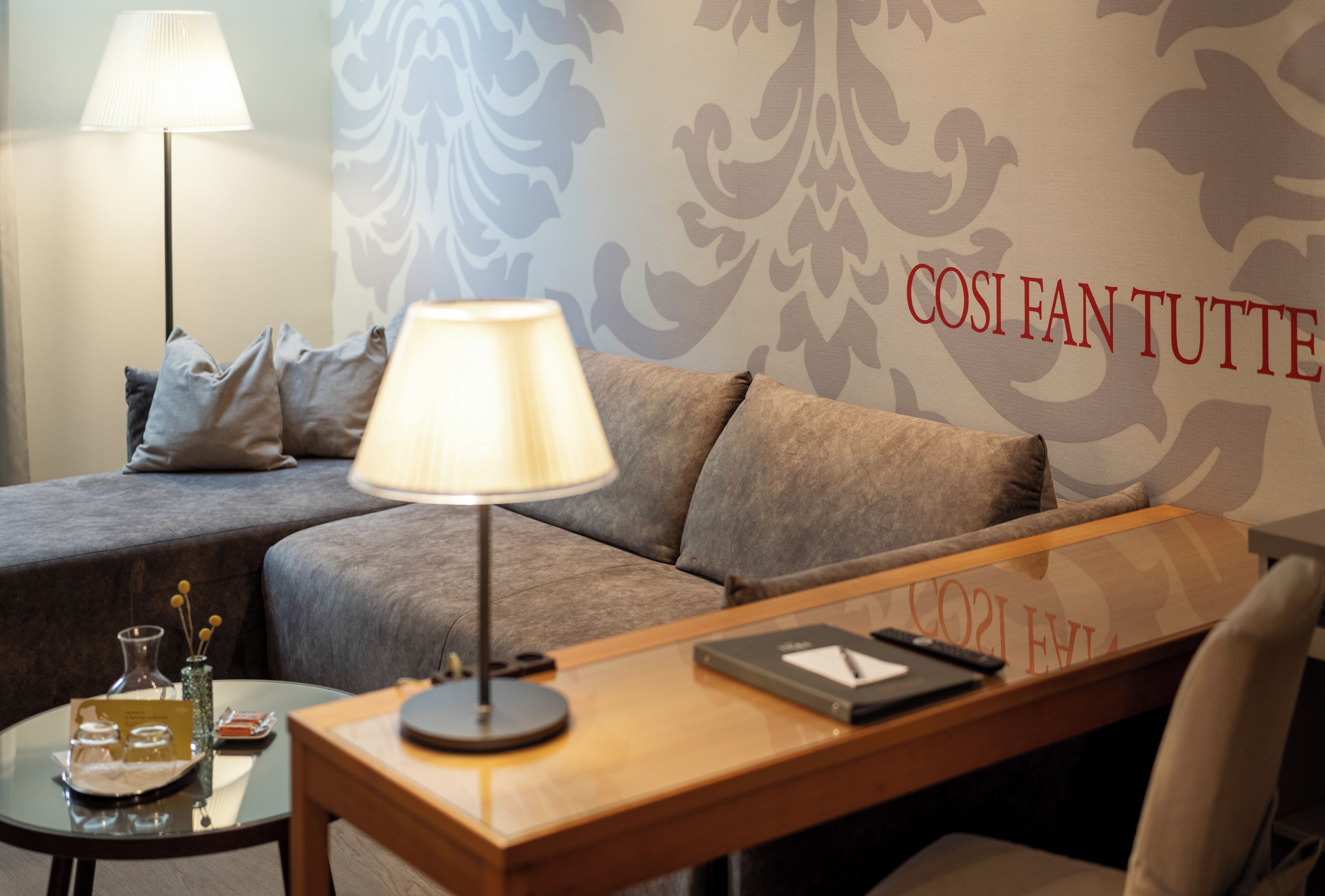 Quarto de hotel com sofá, mesa lateral com abajur, bandeja com copos e parede com padrão decorativo e texto.