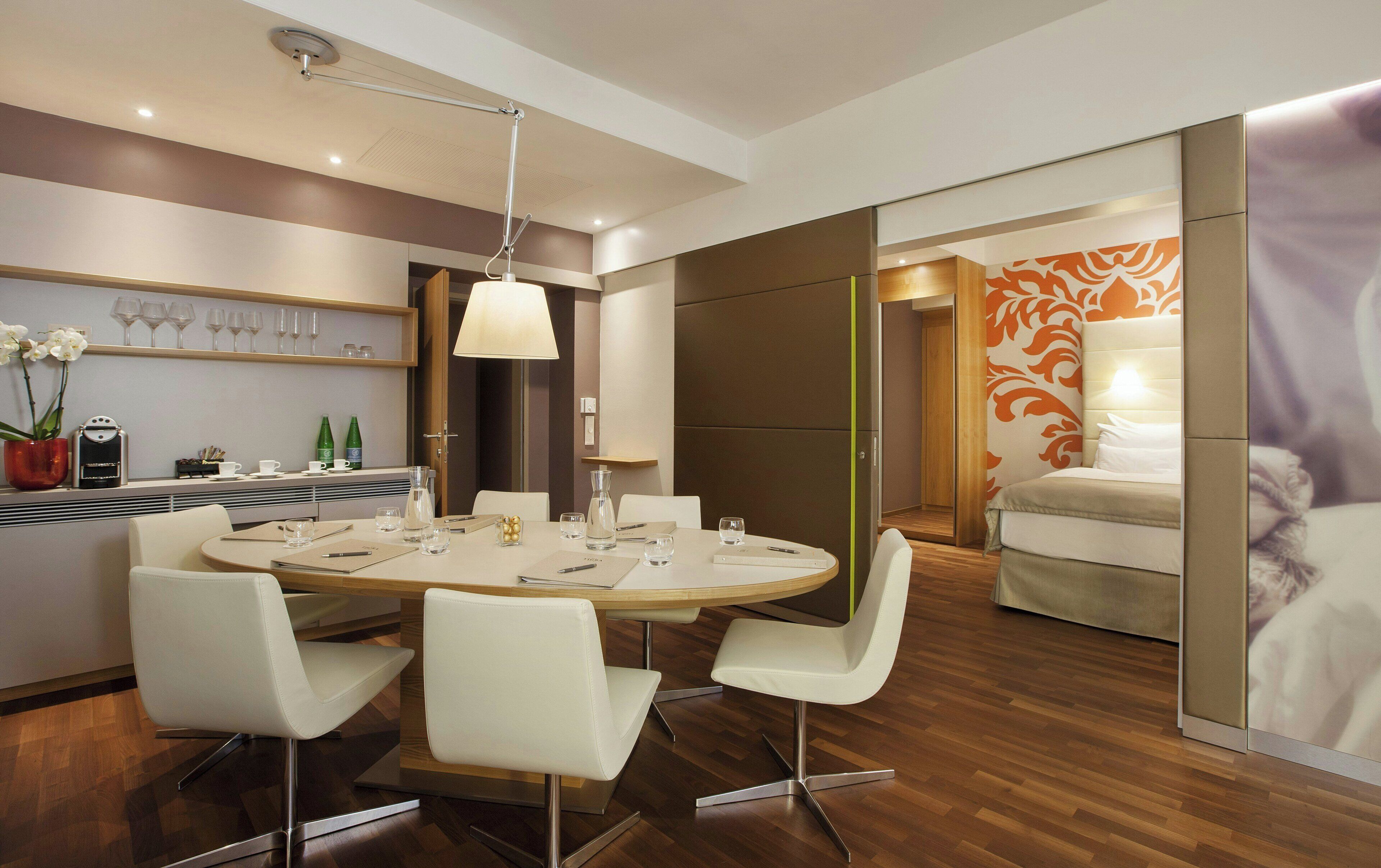 A modern hotel room with a round dining table, white chairs, a coffee machine, and a bed with an orange floral headboard.