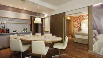 Quarto de hotel moderno com mesa de jantar redonda, cadeiras brancas, máquina de café e cama com cabeceira estampada em flores laranja.