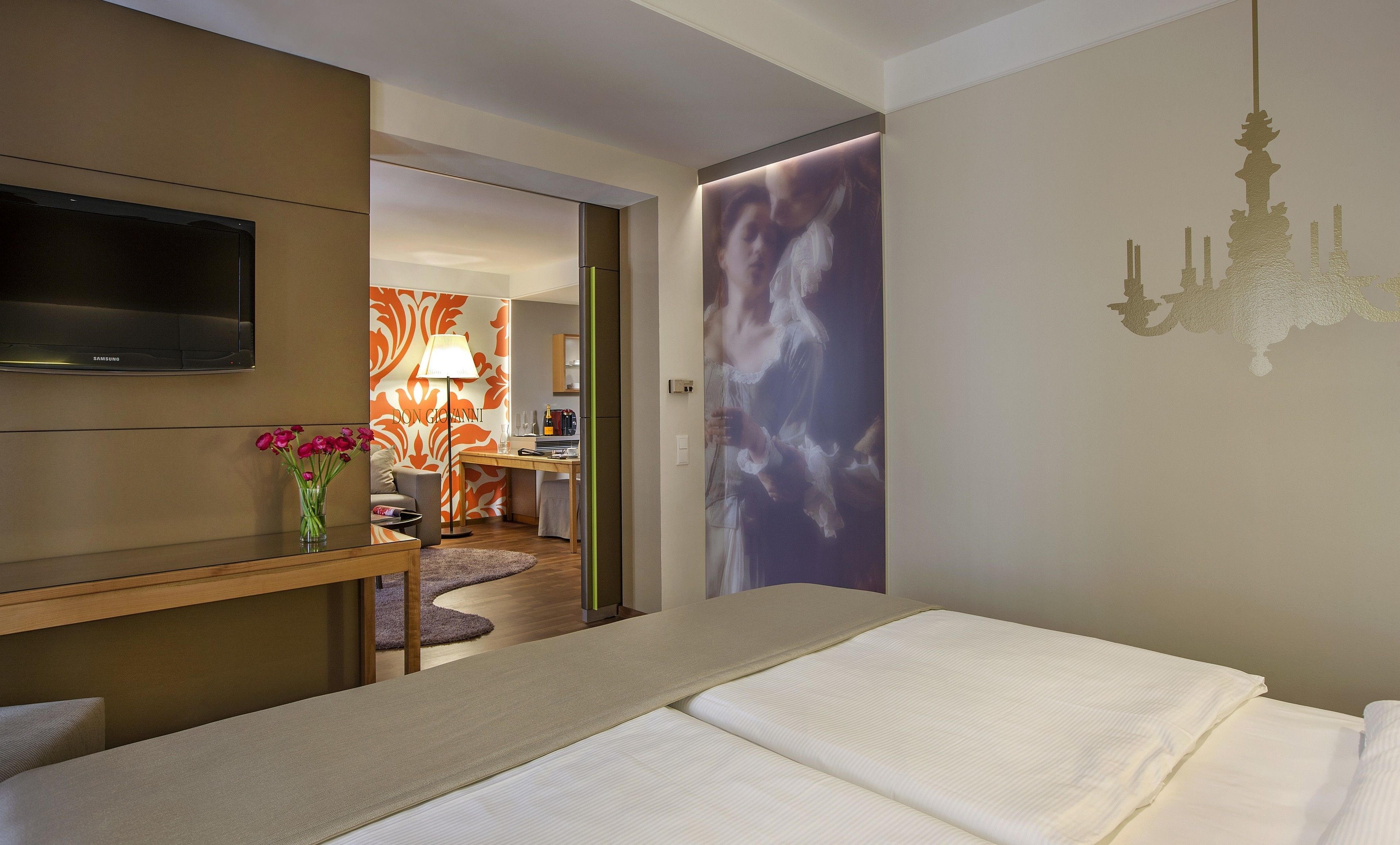 A hotel room with a bed, a TV, a desk with a lamp, and a view of the hallway.