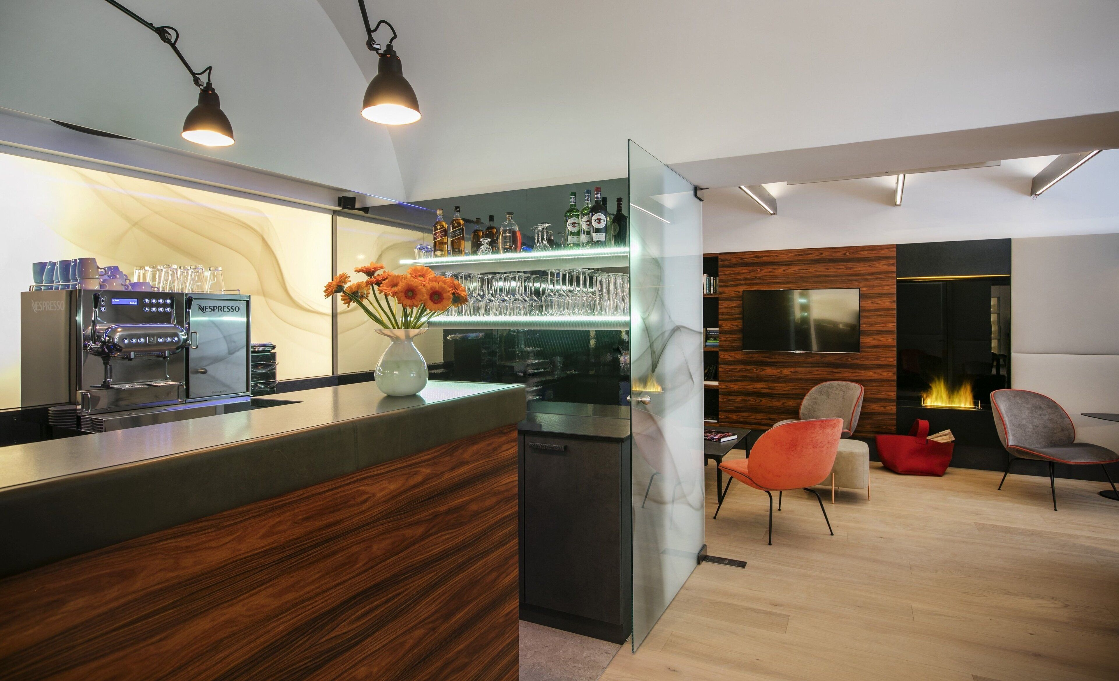 A modern bar area with a Nespresso machine, a glass display case with bottles, and a TV mounted on the wall.