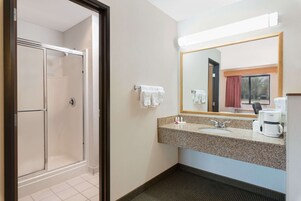 Baño de hotel con cabina de ducha de vidrio, lavamanos con encimera de granito, espejo y cafetera.