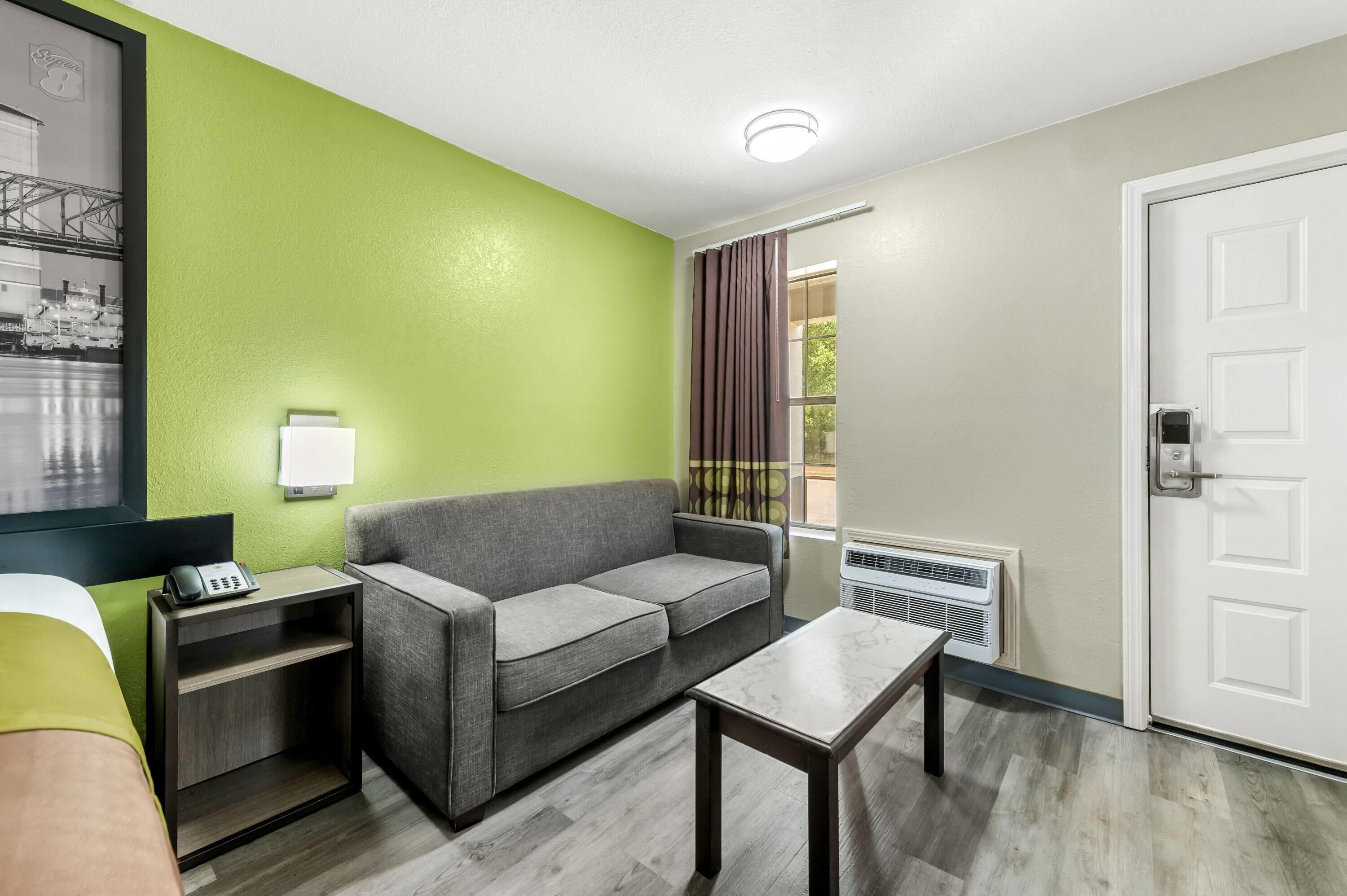Quarto de hotel com sofá cinzento, uma mesinha, luminária, aquecedor e porta.