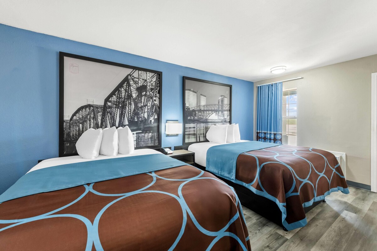 Una camera d'albergo con due letti, un'ampia finestra con tende e opere d'arte alle pareti.
