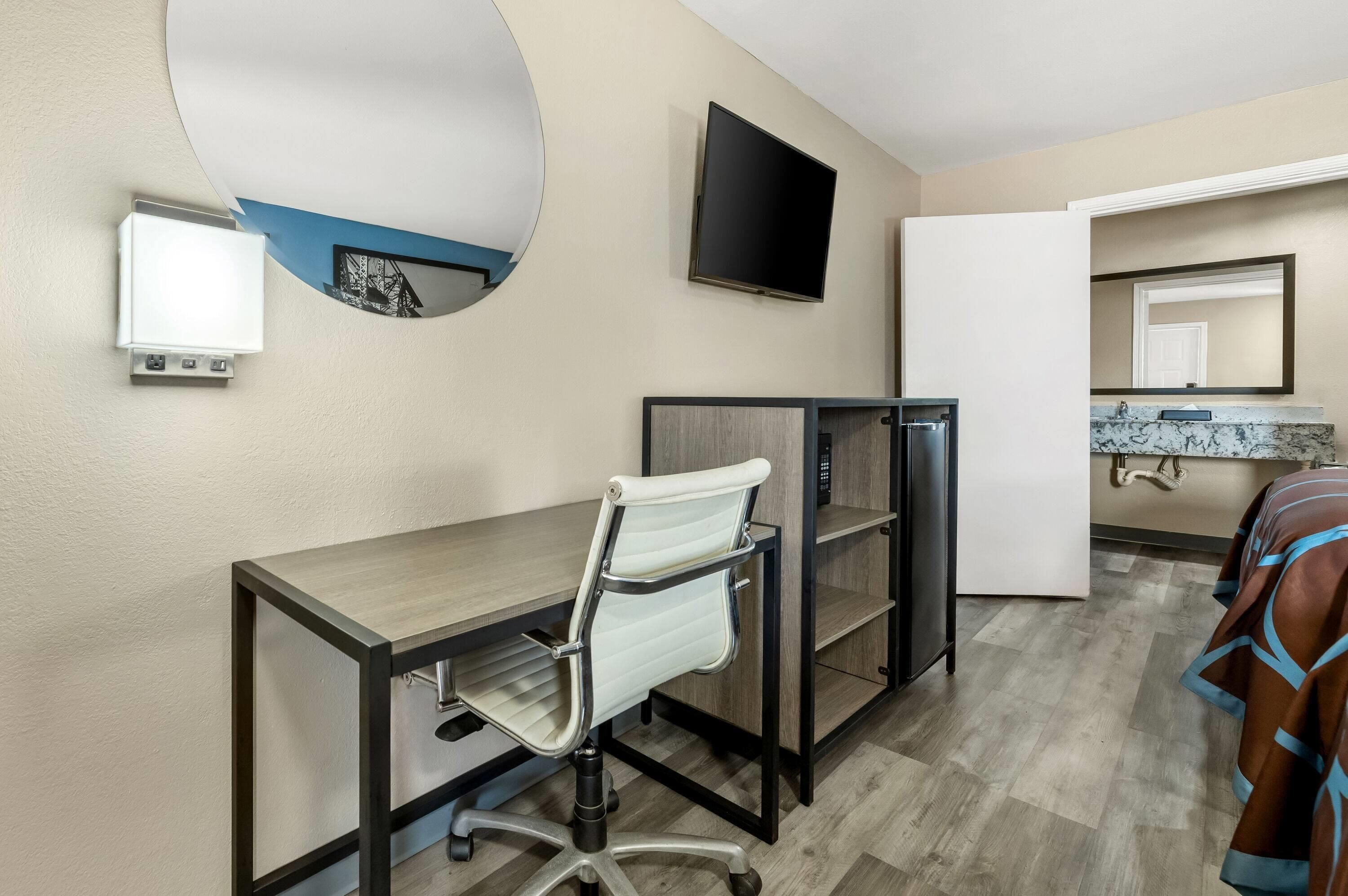 A hotel room with a desk, chair, TV, and a bathroom with a mirror and sink.