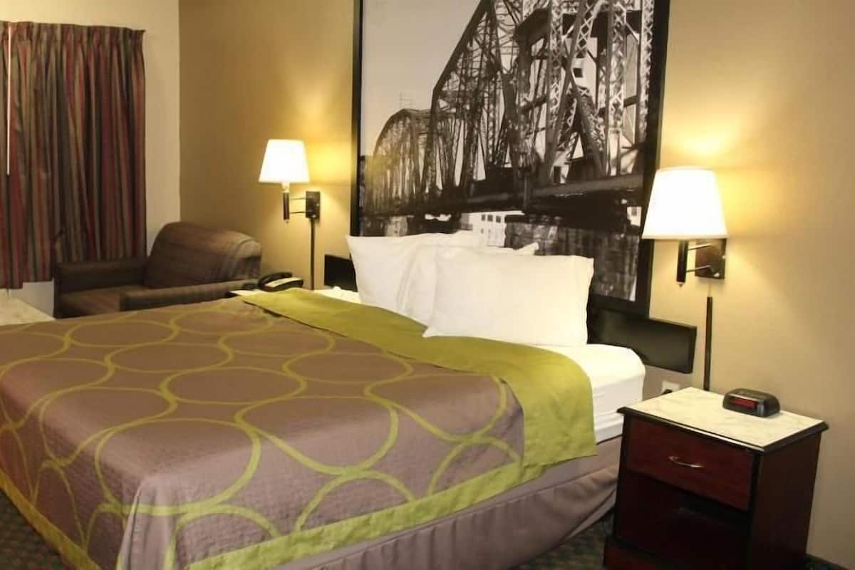 A hotel room with a bed, two bedside lamps, a chair, a desk, and a large painting on the wall.