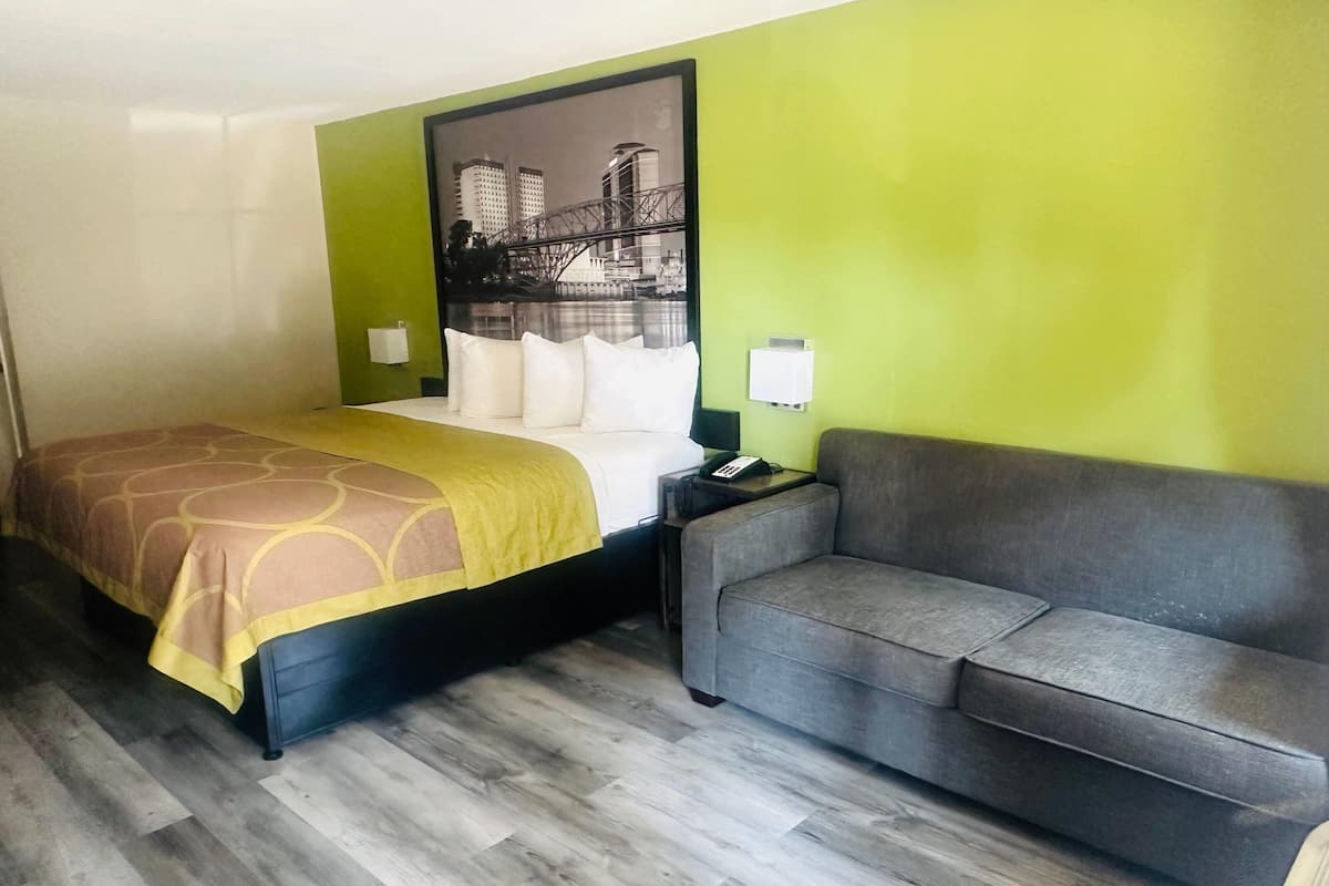 A hotel room with a bed, a grey sofa, a green wall, and a cityscape painting.