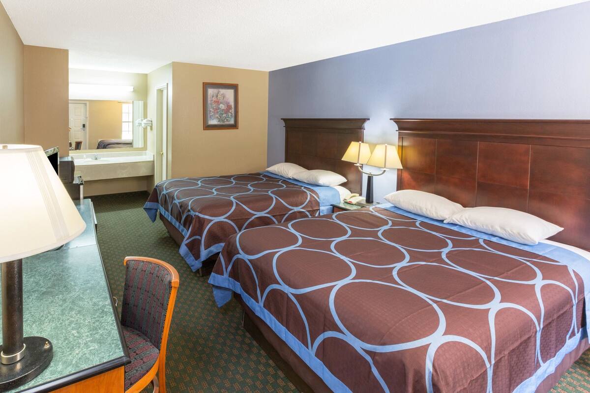 A hotel room with two beds, a desk with a lamp, a chair, and a bathroom visible in the background.