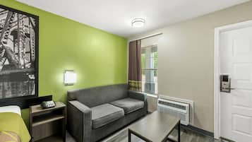 Ein Hotelzimmer mit einer grünen Akzentwand, einem grauen Sofa, einem kleinen Tisch, einem Nachttisch mit Lampe, einem Fenster mit Vorhängen und einer Tür mit Griff.