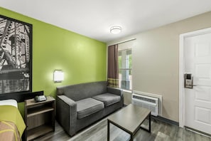 Quarto de hotel com parede verde, sofá cinza, mesa pequena, mesa de cabeceira com abajur, janela com cortinas e porta com puxador.