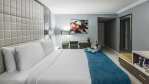 A modern hotel room with a large bed, a flat-screen TV, a desk with a lamp, and a minibar. - Westminster Hotel (Livingston)