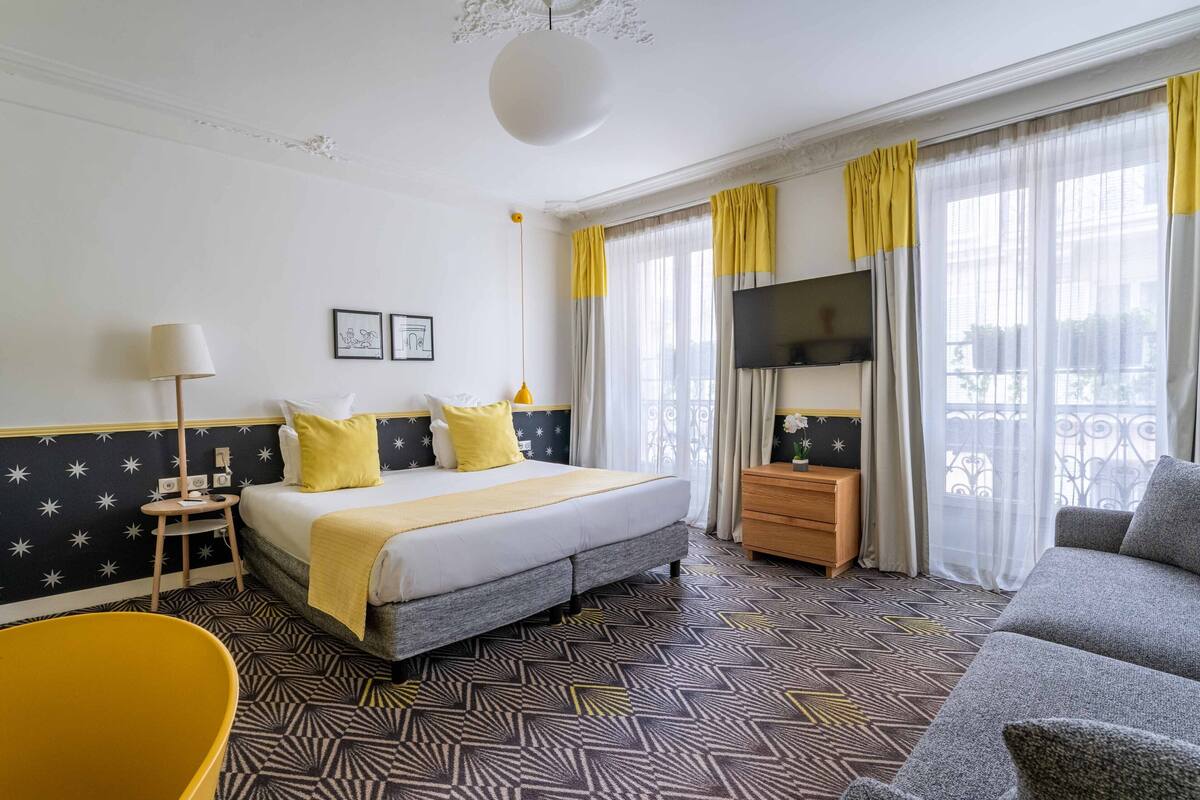 A hotel room with a bed, a sofa, a TV, and a balcony with yellow curtains.