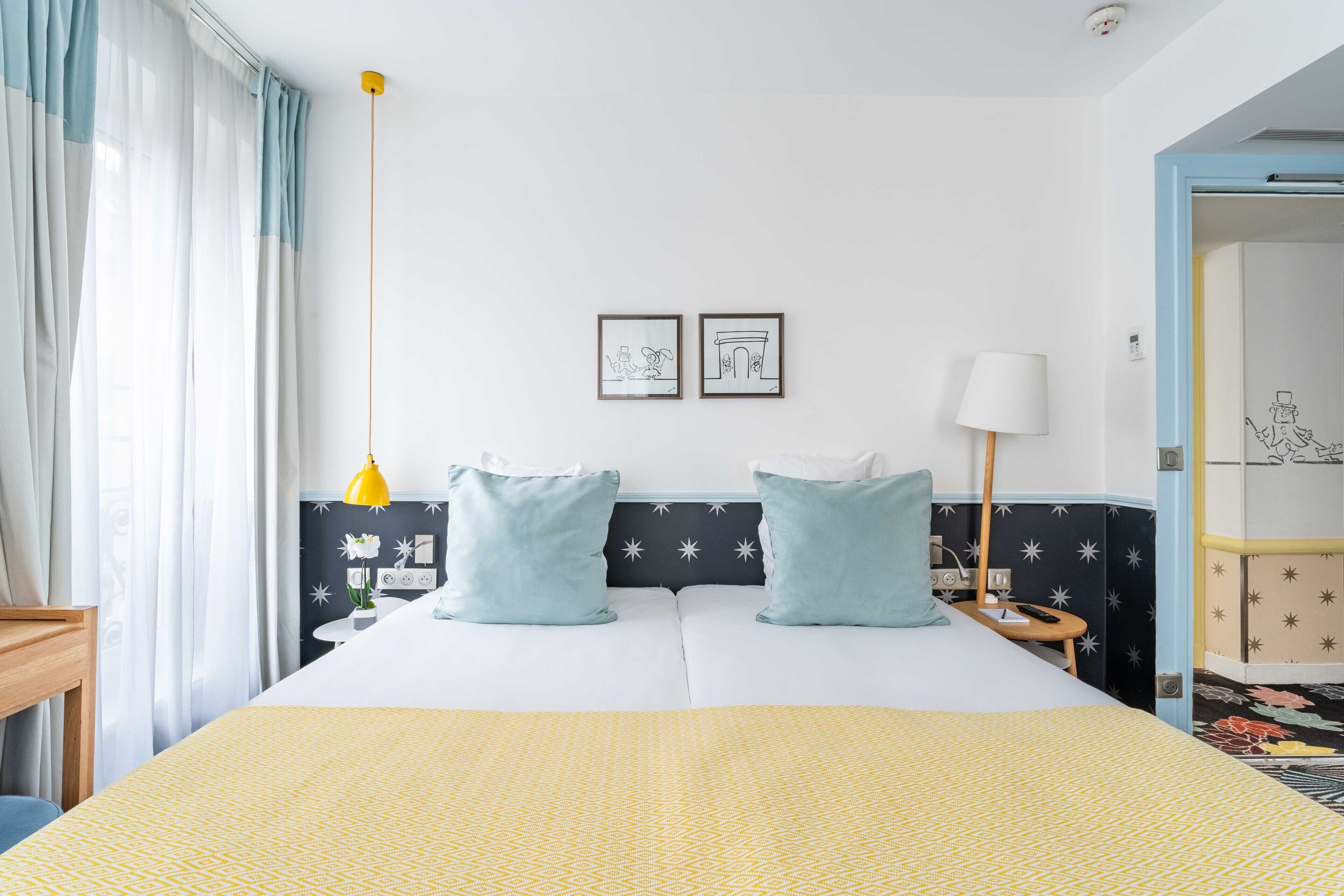 Une chambre d’hôtel avec un lit, deux oreillers, une table de chevet, une lampe et un mur orné de deux tableaux encadrés.