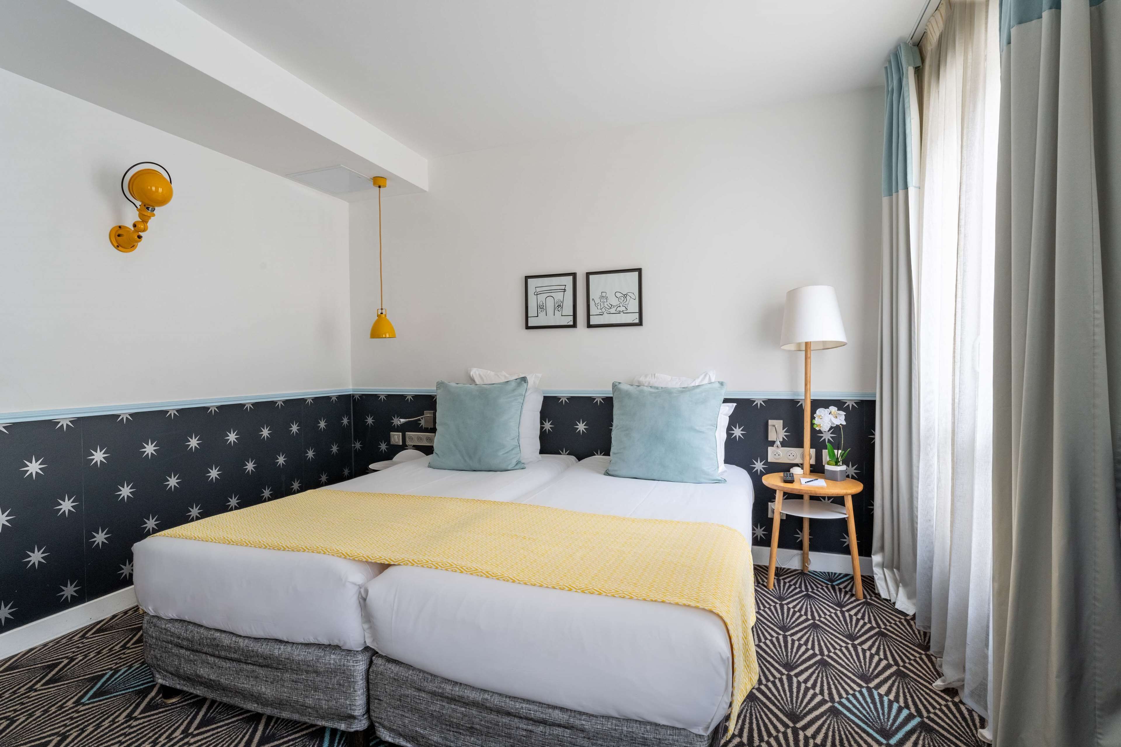 A hotel room with a bed, a bedside table with a lamp, a small table with a plant, and star-patterned walls.