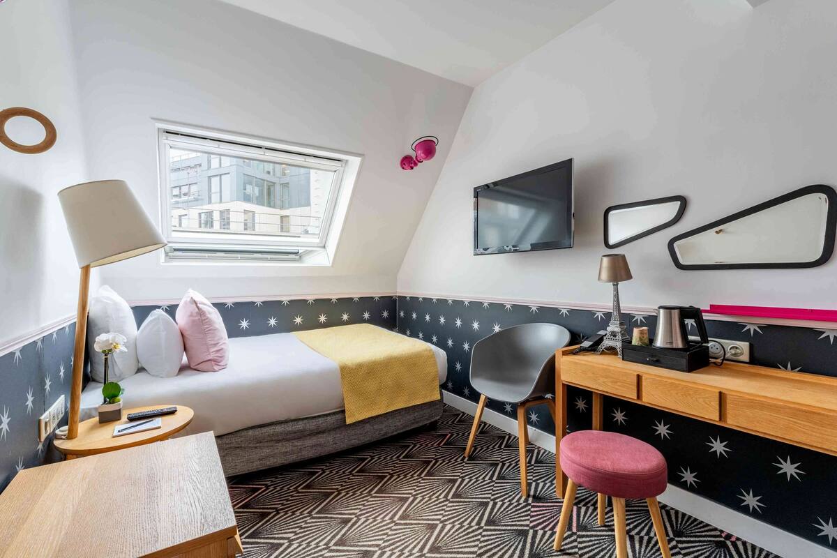 A compact hotel room with a bed, a desk, a chair, a small table, a TV, and a window with a view of buildings.
