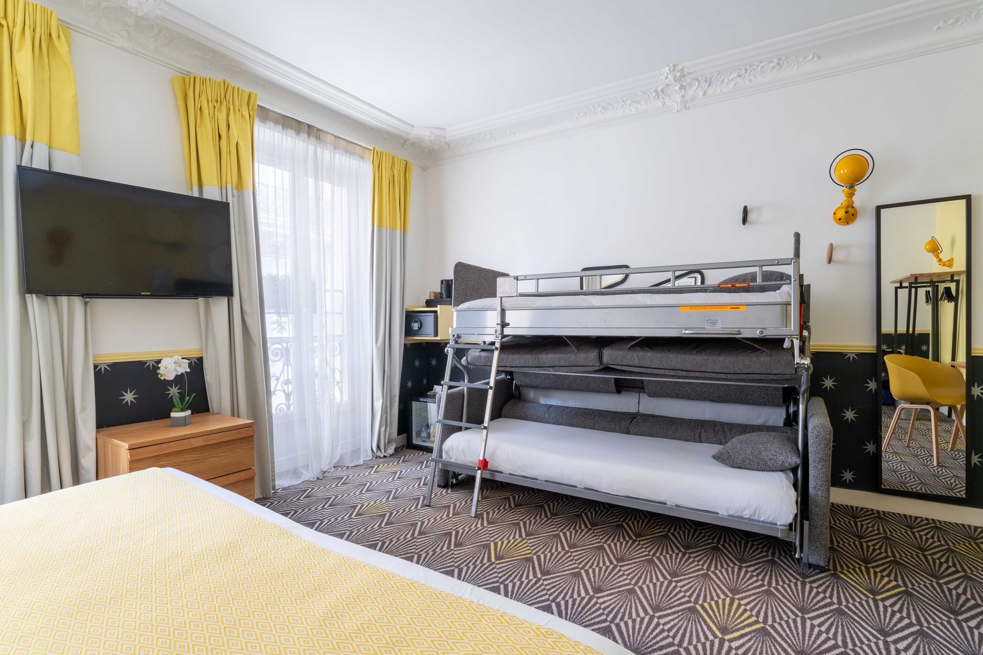 A hotel room with a bunk bed, a flat-screen TV mounted on the wall, a wooden nightstand with a small table, and a mirror with a yellow frame.