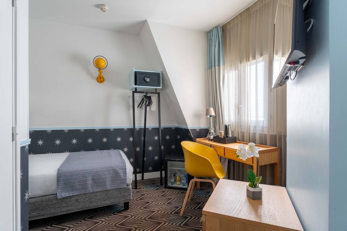 A bedroom with a bed, a desk, a chair, a small table with a plant, a wall-mounted lamp, a key holder, and a thermostat.