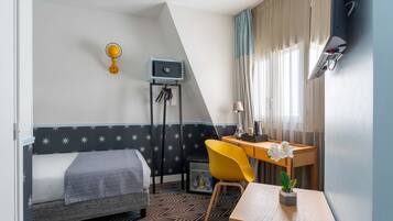 A bedroom with a bed, a desk, a chair, a small table with a plant, a wall-mounted lamp, a key holder, and a thermostat.