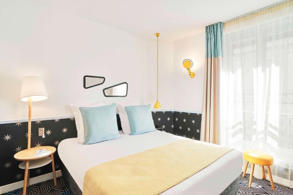 A hotel room with a bed, a bedside table, a lamp, a stool, and a window with curtains.