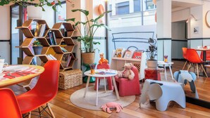 A cozy, well-lit room with a wooden floor, a round table with a red chair, a bookshelf, and a window with a view of buildings. - Hôtel Augustin - Astotel (Paris)