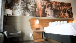 Ett hotellrum med en säng, en stol, ett nattduksbord med en lampa och en stor väggmålning med ett grottmotiv.