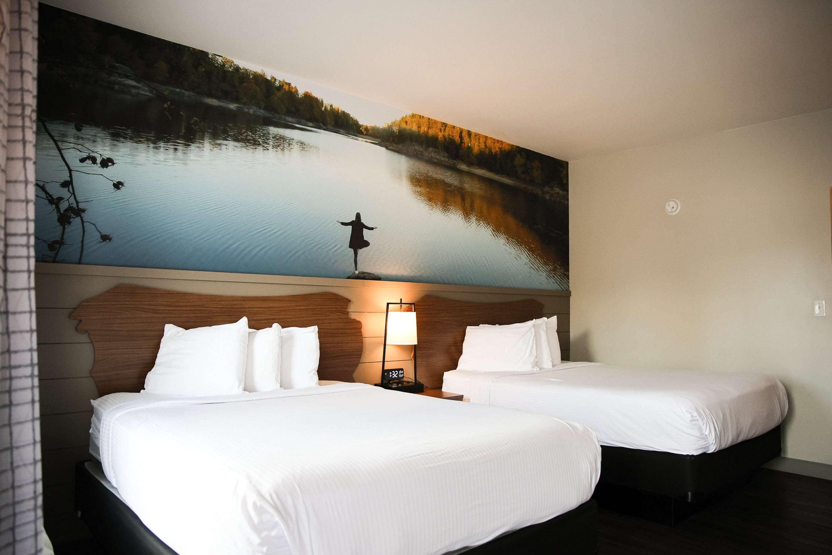 Habitación de hotel con dos camas, un cuadro grande de un lago y una mesita de noche con lámpara.