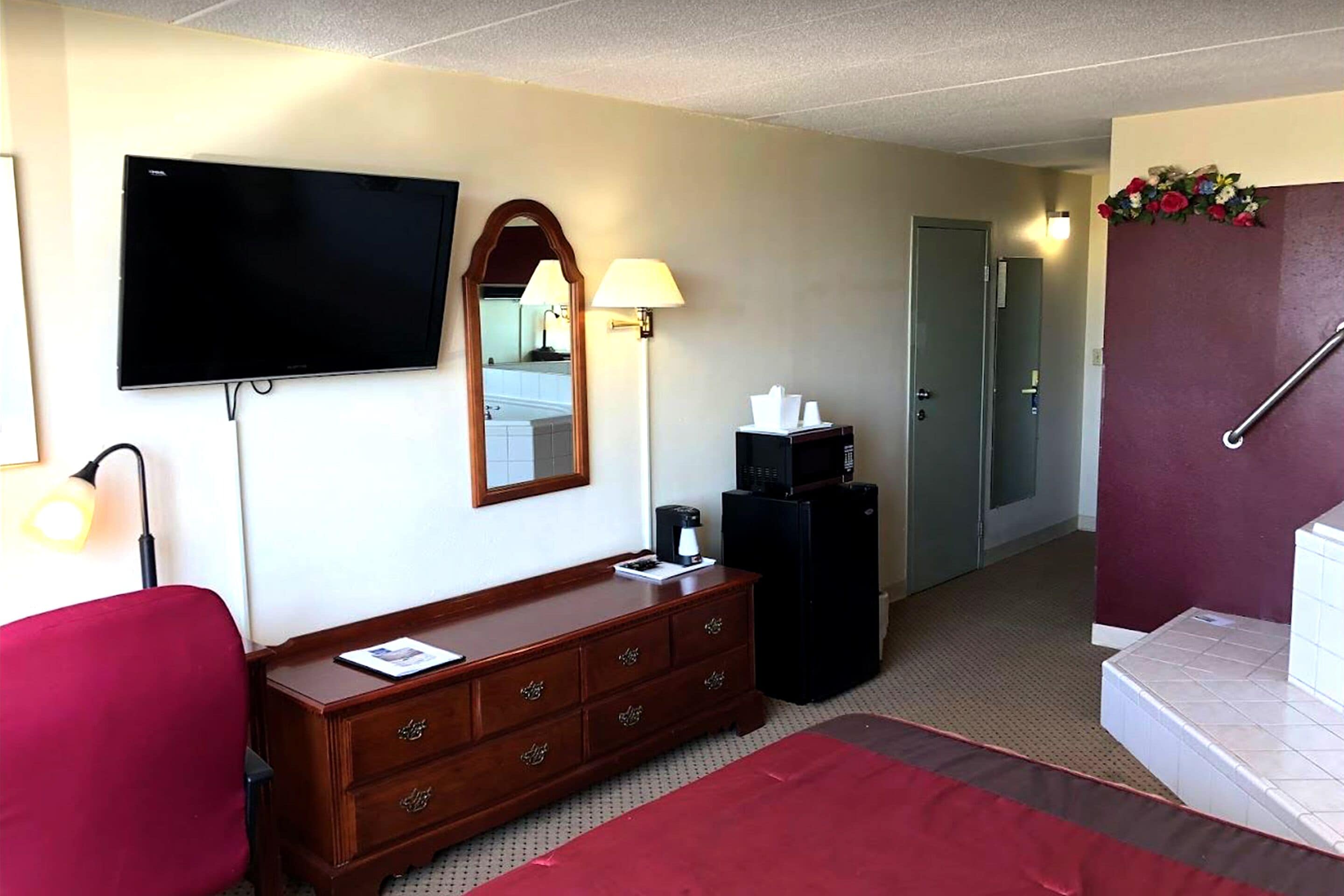 A hotel room with a bed, a wooden dresser, a television, a mirror, a desk, and a desk lamp.