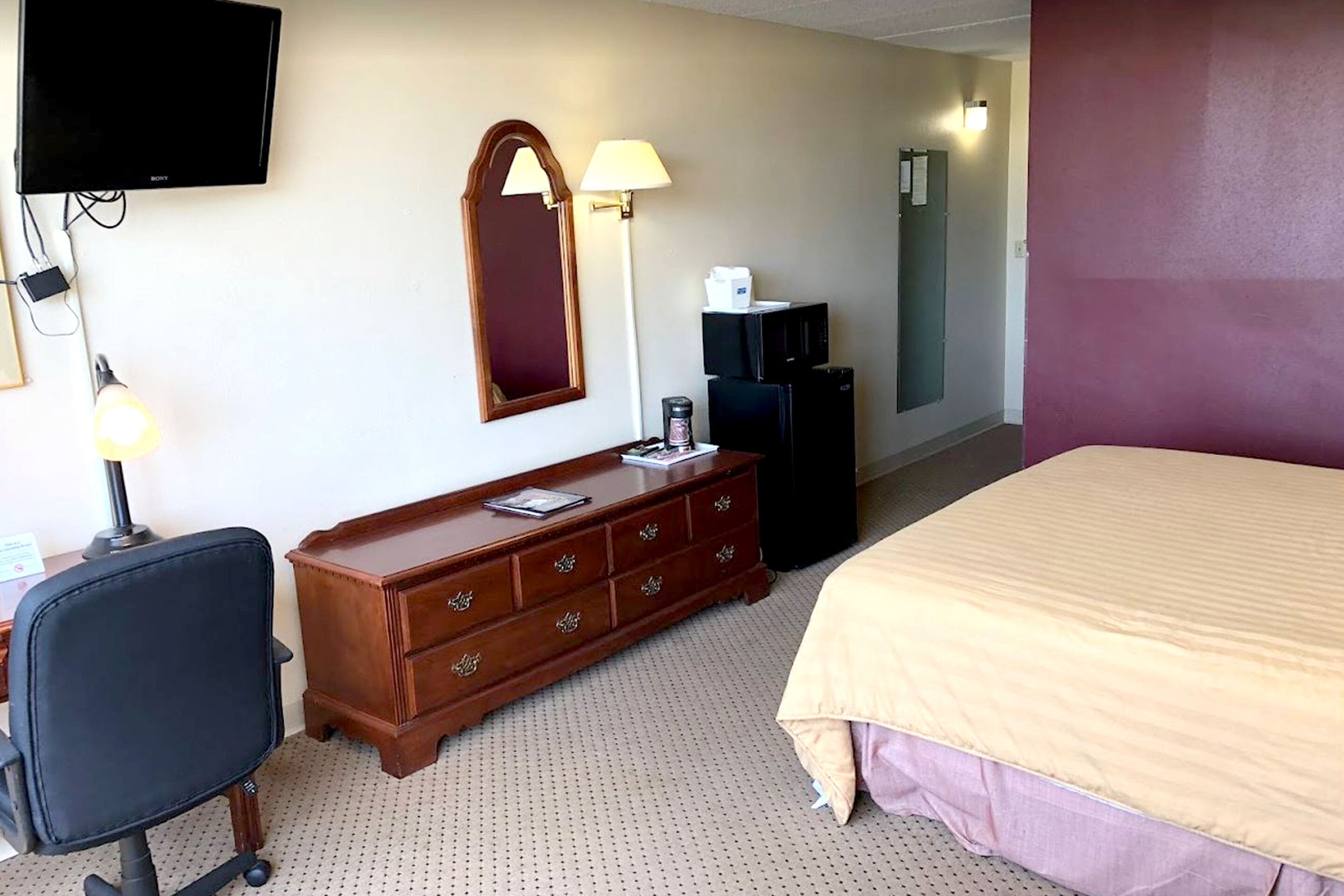 Ein Hotelzimmer mit einem Bett, einem Schreibtisch mit Stuhl, einem Schminktisch, einem Fernseher, einer Lampe und einem Kühlschrank.