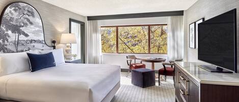 A hotel room with a large bed, a desk, a chair, a television, and a window with a view of trees.