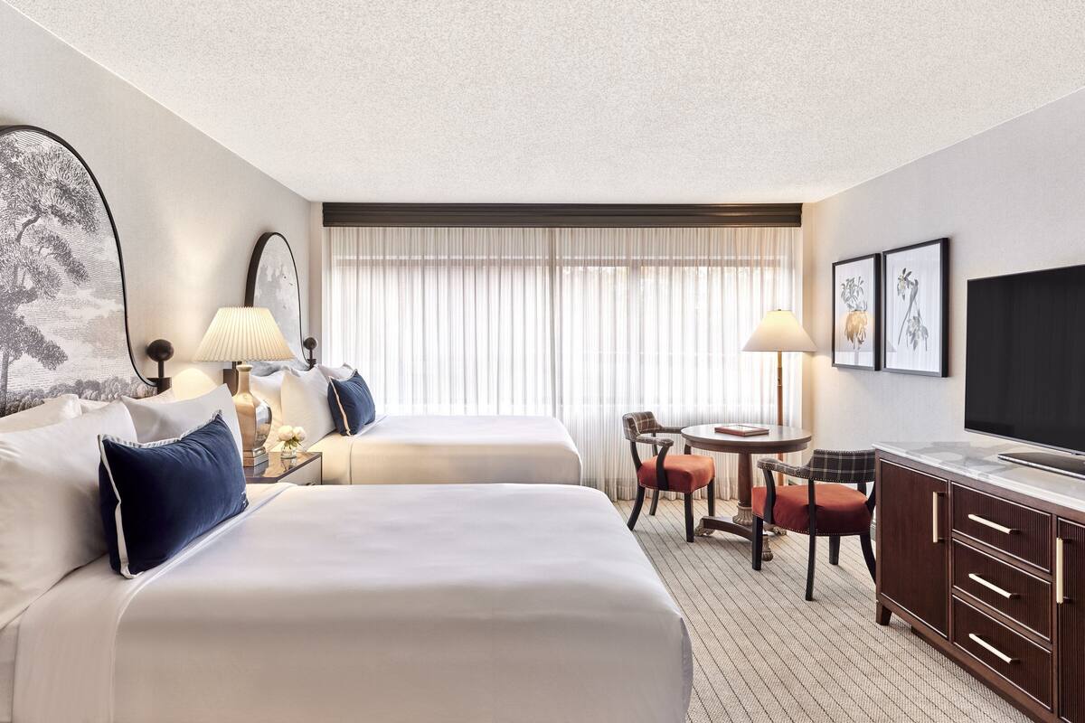 A hotel room with two beds, a desk with a chair, a television, and a window with curtains.