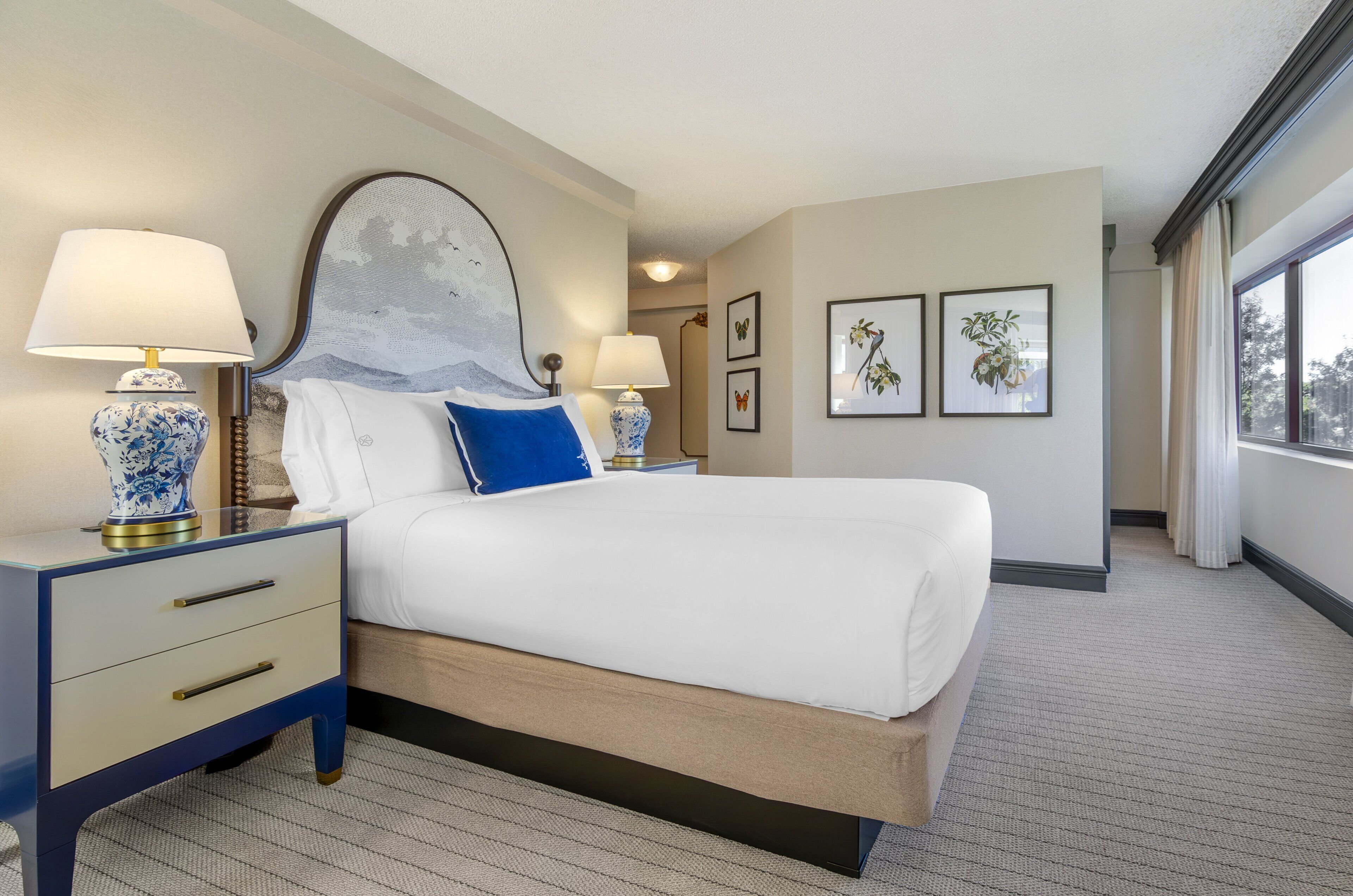 Una habitación de hotel con una cama grande, dos mesitas de noche con lámparas, una mesita auxiliar y cuadros enmarcados en la pared.