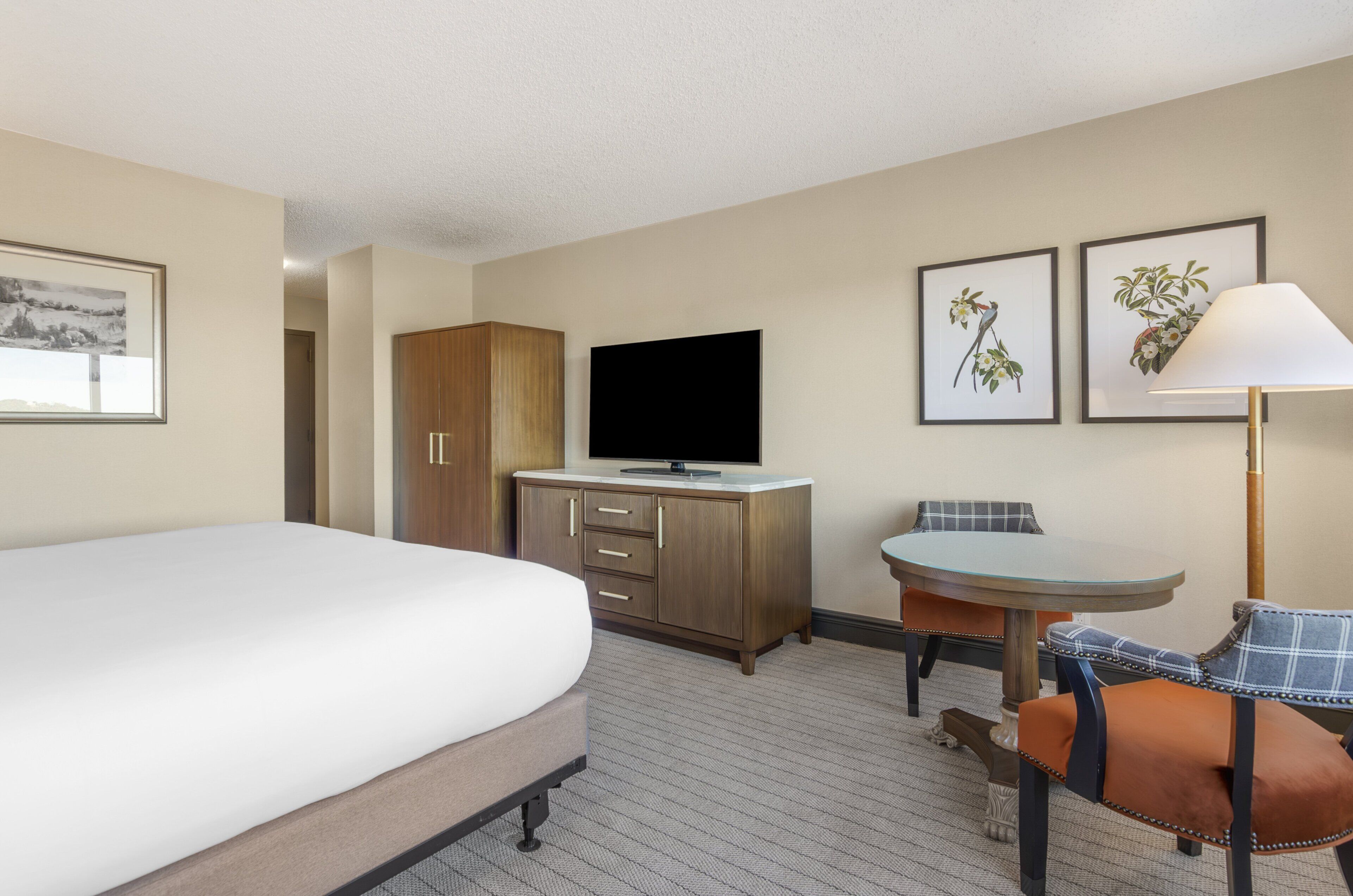 Une chambre d’hôtel avec un lit, un téléviseur posé sur une commode en bois, une petite table avec une chaise, et deux tableaux encadrés accrochés au mur.