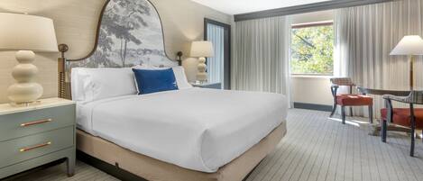 A hotel room with a large bed, two bedside tables with lamps, a desk with a chair, and a window with curtains.