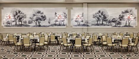 A banquet hall with round tables and chairs, large windows, and a geometric patterned carpet.