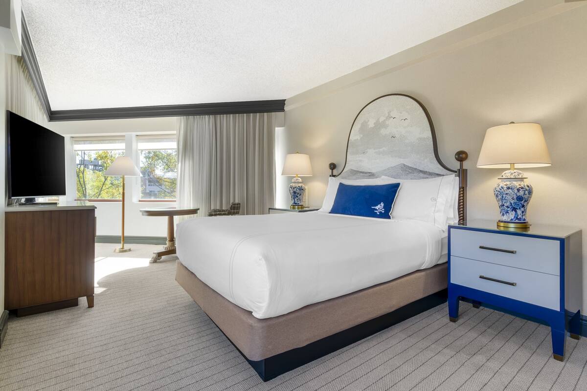 A hotel room with a large bed, two bedside tables with lamps, a flat-screen TV, a wooden dresser, and a view of the outdoors.