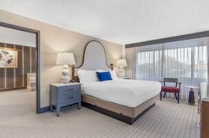 Una habitación de hotel con una cama grande, dos mesitas de noche con lámparas, una silla, una pequeña mesa y un espejo grande.