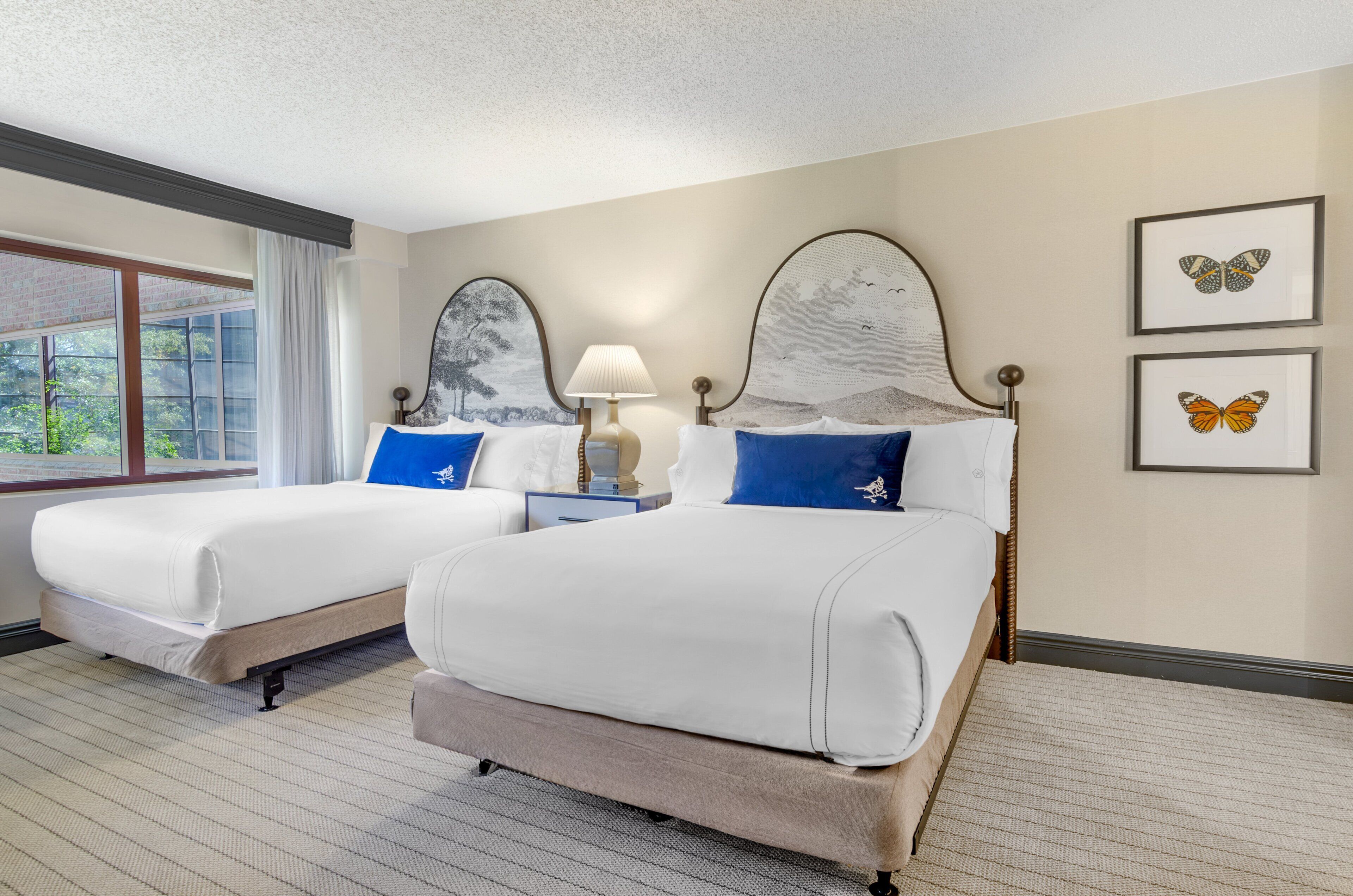 Habitación de hotel con dos camas, una mesita de noche, una lámpara y cuadros con mariposas en la pared.