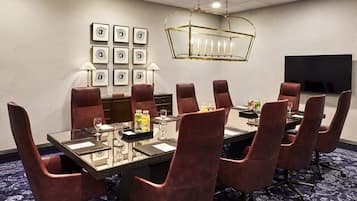 Une salle de conférence avec une table en verre, des chaises à revêtement rouge, un lustre, des tableaux encadrés au mur et un téléviseur à écran plat.