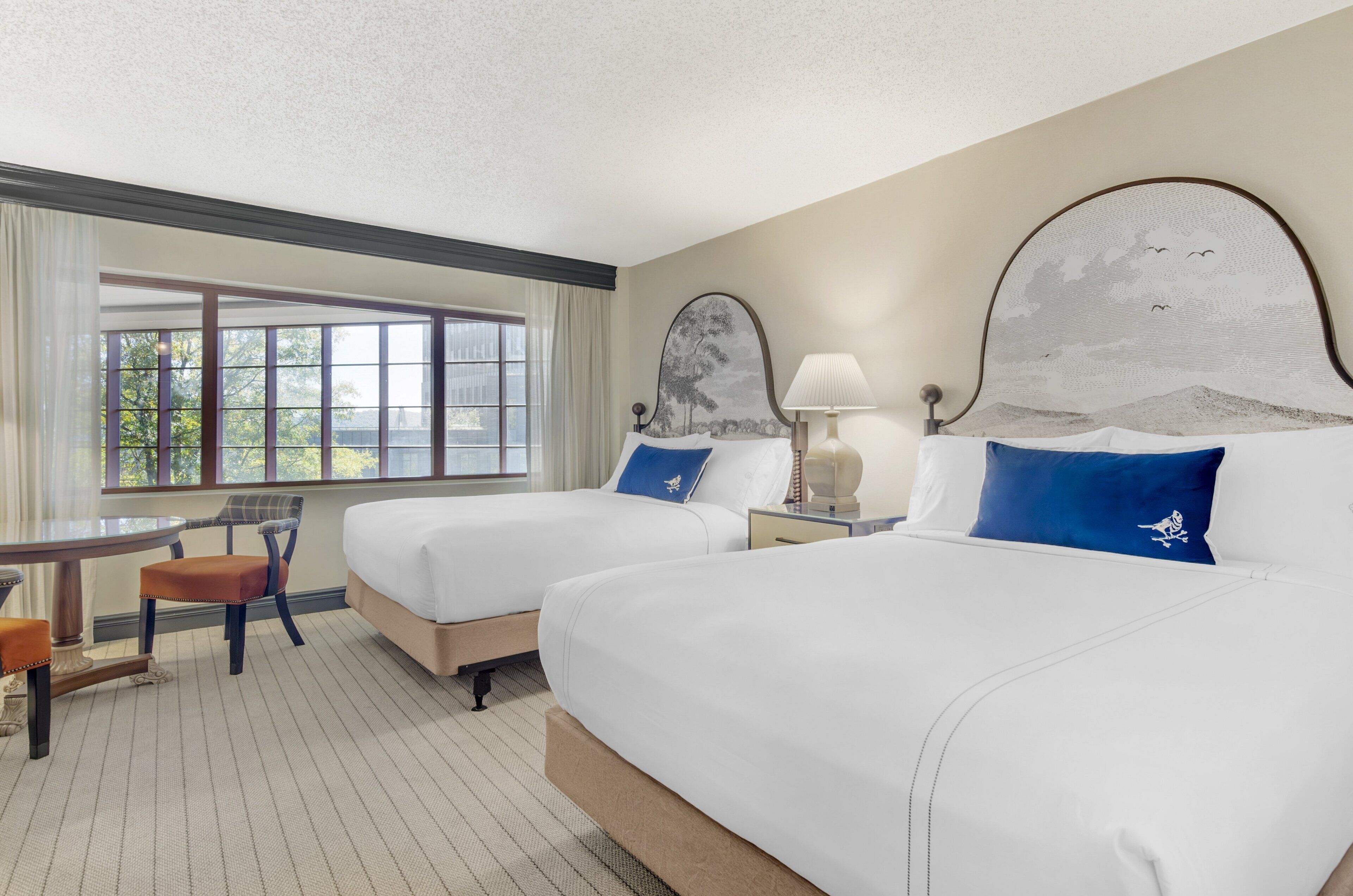 A hotel room with two beds, a desk with a chair, and a large window.