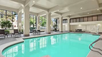 An indoor swimming pool with clear views of the outdoors, a seating area with tables and chairs, and a staircase leading to an upper level.