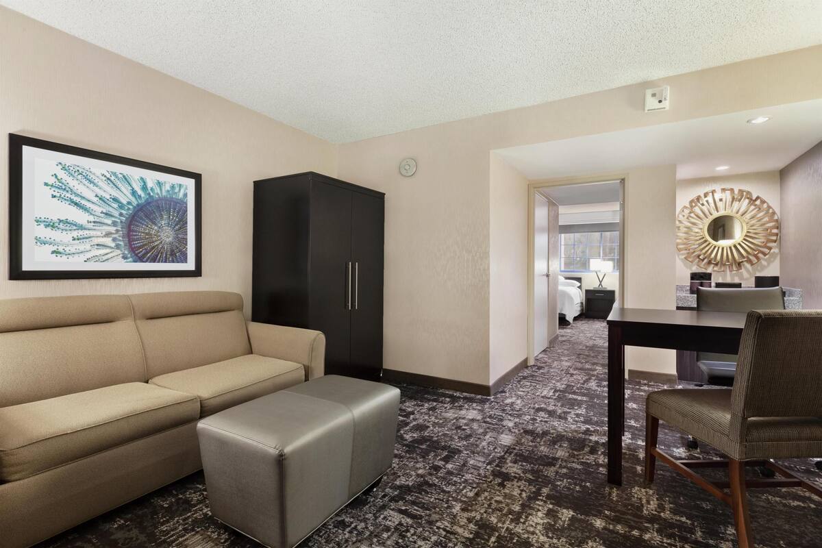 Quarto de hotel com sofá bege, um banquinho cinza, guarda-roupa preto, mesa de jantar com uma cadeira e um espelho decorativo.