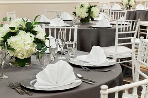 A table set with white napkins, silverware, and clear glasses, surrounded by white chairs and a vase of white flowers.
