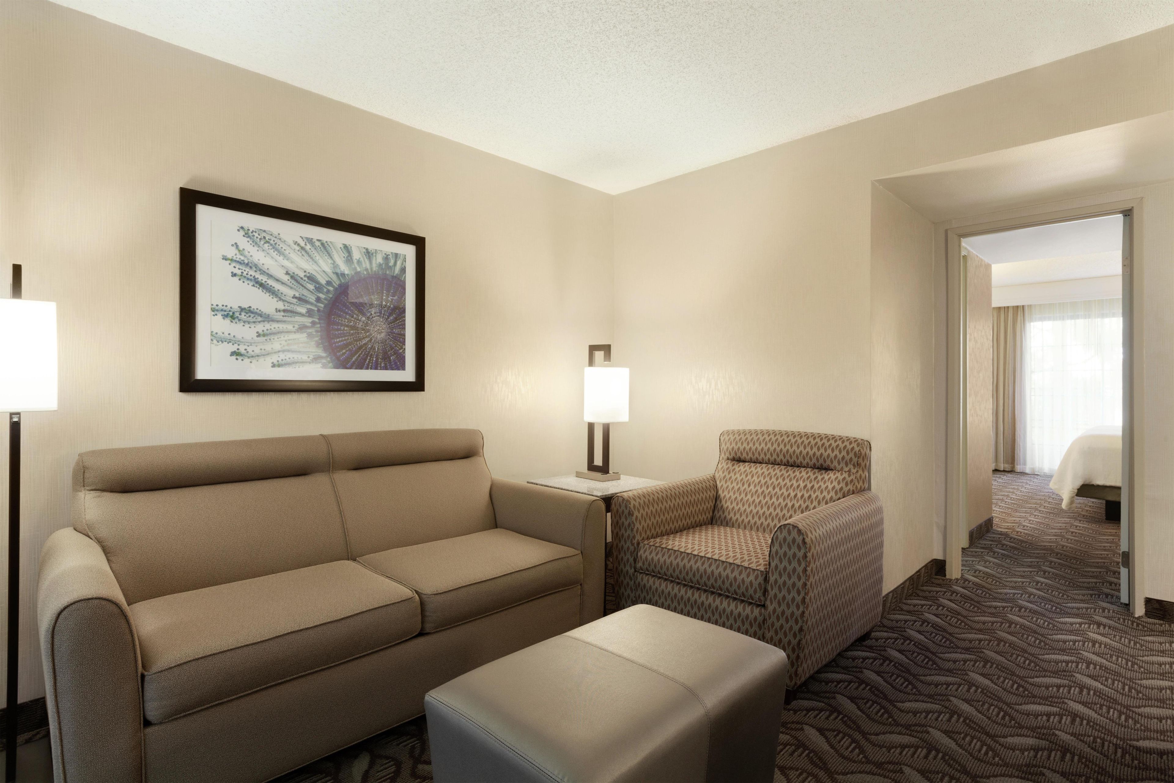 A hotel room with a sofa, armchair, and ottoman, a framed artwork on the wall, and a patterned carpet.