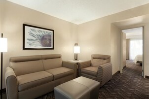A hotel room with a sofa, armchair, and ottoman, a framed artwork on the wall, and a patterned carpet.