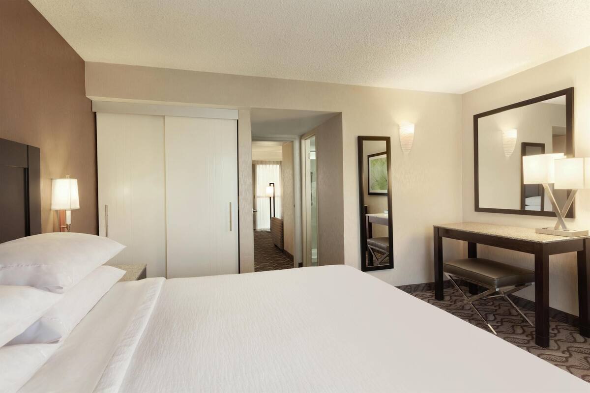Quarto de hotel com uma cama, escrivaninha, cadeira, espelho e porta que dá para um corredor.
