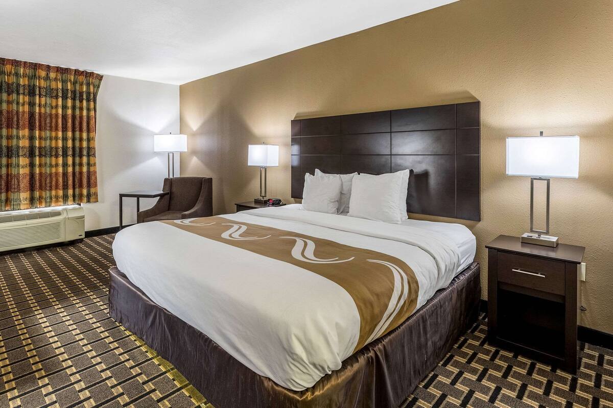 A hotel room with a large bed, two bedside tables with lamps, a chair, and a patterned carpet.
