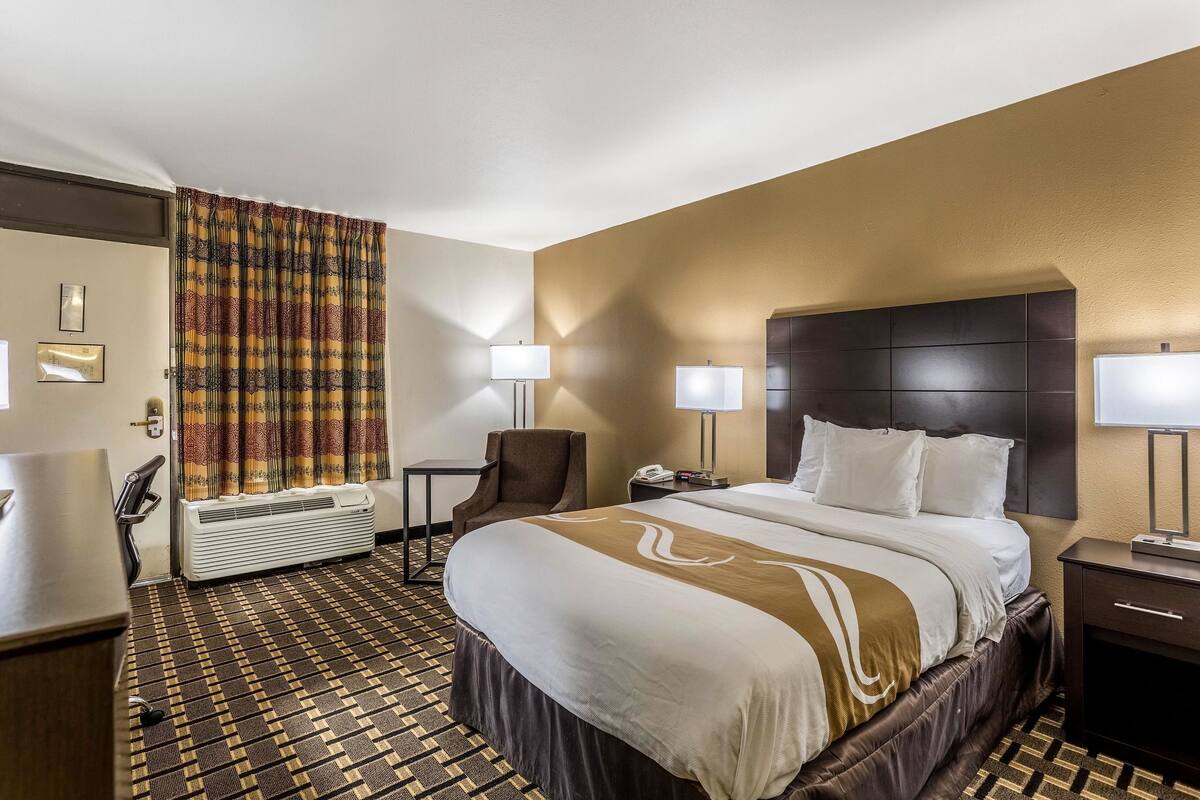 A hotel room with a large bed, two bedside tables, a chair, an air conditioning unit, and a curtain.