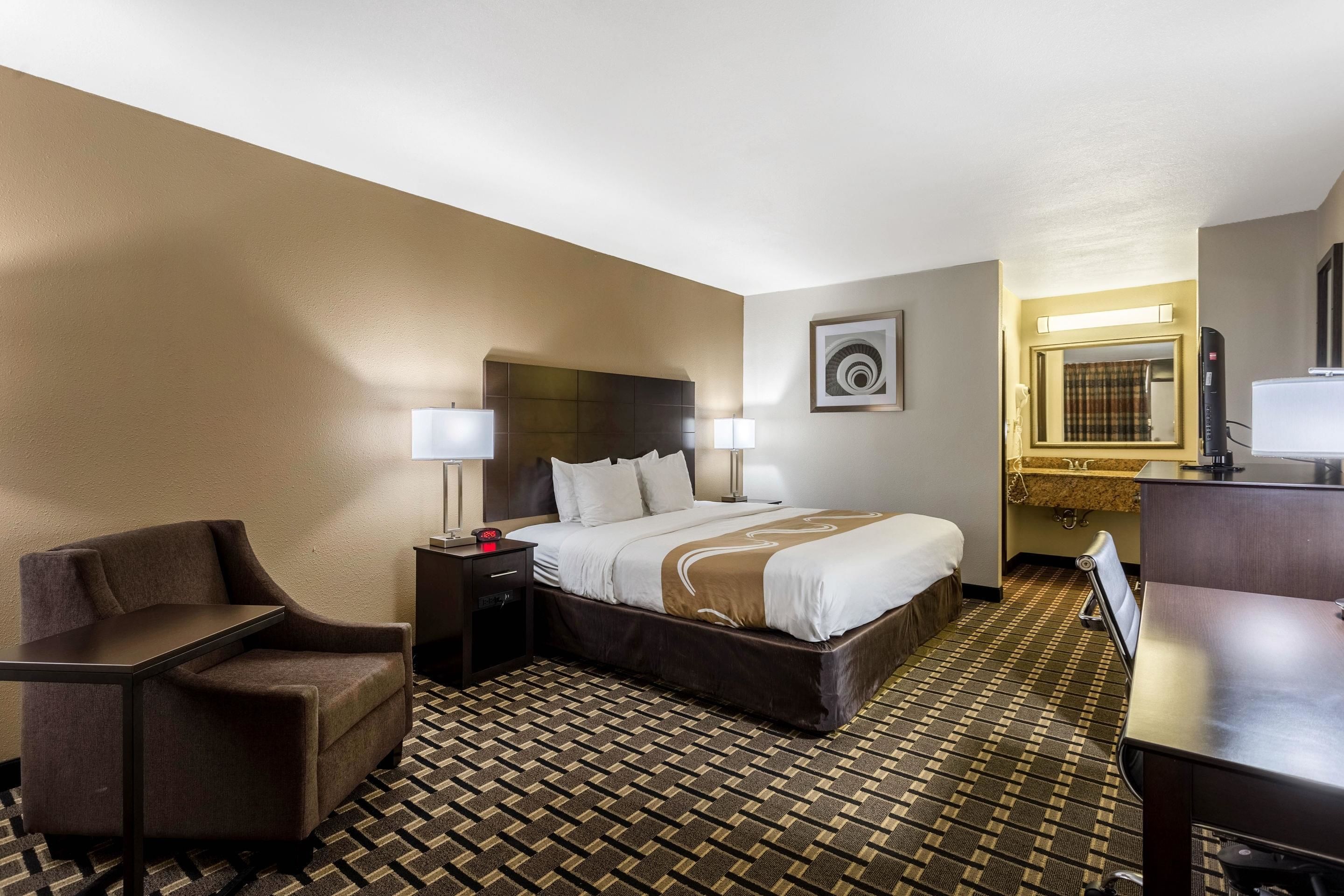 Habitación de hotel con cama, escritorio, silla, mesita de noche, lámpara y espejo de baño.